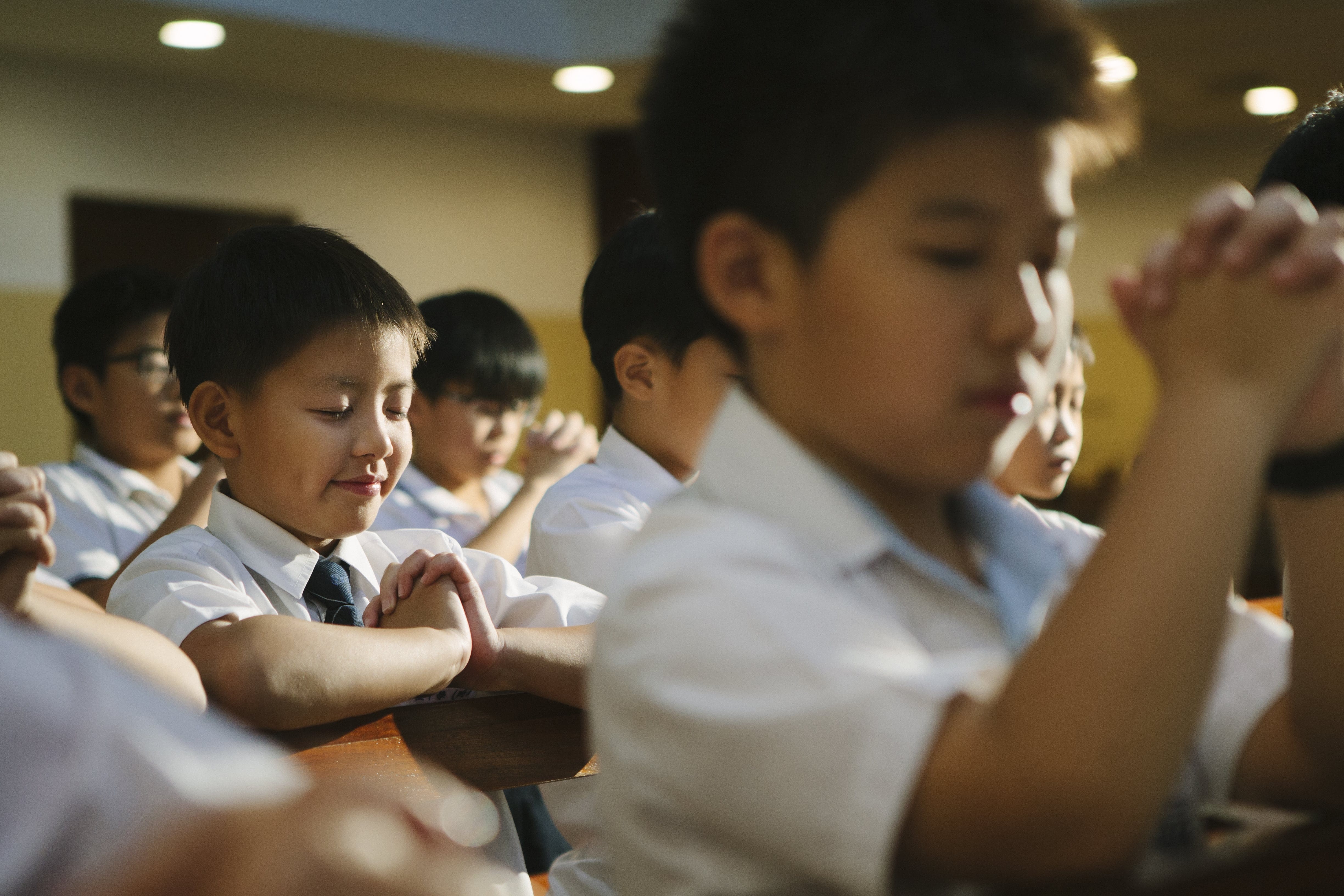Several young boys in white school uniforms sit with hands clasped and eyes closed, praying in a chapel.